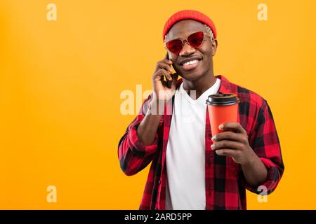 Gutaussehender schwarzer amerikaner, der auf dem Telefon spricht, während er ein wegnehmendes Kaffee-Glas auf gelbem Studiohintergrund mit Kopierbereich hält. Stockfoto