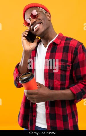 Stilvoller schwarzer amerikanischer Mann mit einem schönen Lächeln in einem karierten roten Hemd hält ein Glas Kaffee auf gelbem Studiohintergrund in seinen Händen. Stockfoto