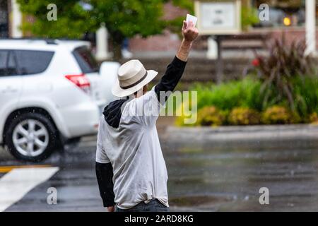 Rückansicht des Mannes, der Hut und weißes T-Shirt trägt, während er weißes Gewebepapier zeigt und eine Ankündigung auf der Straße macht. In der Regenzeit Stockfoto