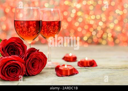 Valentinstag Konzept. Zwei Gläser Wein, rote Rosen und brennende Kerzen auf einem alten Holztisch vor einem verschwommenen leuchtenden Hintergrund mit Kopie Stockfoto