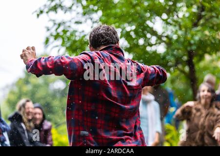 Rückansicht eines unbeschwerten Mannes, der in Rot und Blau kariertes Hemd trägt, während er tanzt und während der sommerlichen Regenzeit gegen Menschen in der Stadt genießt Stockfoto