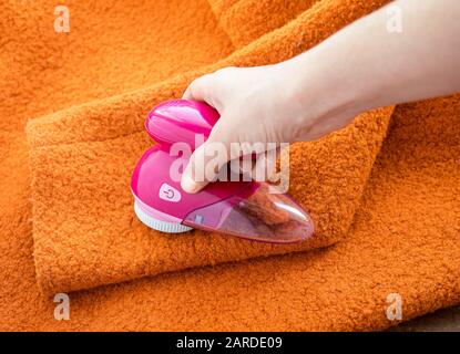 Pflege der Kleidung. Flusenrasierer oder Stoffrasierer oder Fuzz-Entferner in weiblicher Hand. Frau entfernt Flusen auf orangefarbenem Wollmantel mit elektrischem Entfussler. Stockfoto
