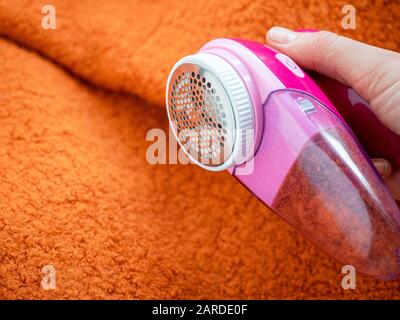 Pflege der Kleidung. Flusenrasierer oder Stoffrasierer oder Fuzz-Entferner in weiblicher Hand. Frau entfernt Flusen auf orangefarbenem Wollmantel mit elektrischem Entfussler. Stockfoto