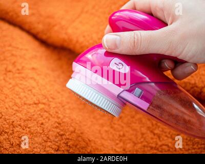 Pflege der Kleidung. Flusenrasierer oder Stoffrasierer oder Fuzz-Entferner in weiblicher Hand. Frau entfernt Flusen auf orangefarbenem Wollmantel mit elektrischem Entfussler. Stockfoto