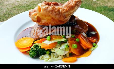 Traditioneller englischer Roast mit Yorkshire Pudding, serviert mit geschnittenem Rindfleisch, Karotten, Erbsen, Brokkoli und Gravy auf einem weißen Teller. Stockfoto