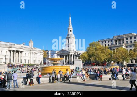 Touristen, die auf einem sonnigen Trafalgar Square mit der St. Martin-in-the-Fields-Kirche im Hintergrund sitzen, London, England, Großbritannien Stockfoto