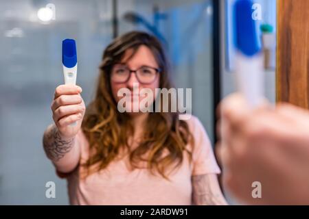 Ein Blick über die Schulter als schöne kaukasische Frau steht vor dem Badezimmerspiegel mit einem Schwangerschaftstest aus Kunststoff, der ein positives Ergebnis zeigt Stockfoto