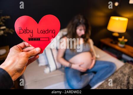 Eine selektive Fokussierung, die konzeptionell aufgenommen wird, wenn zukünftige Eltern sich auf ein valentinskind freuen. Rotes Herz mit Botschaft halten, während die Dame auf Baby-Bump blickt Stockfoto