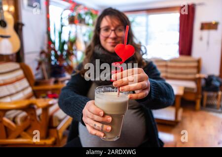 Ein selektiver Fokusschuss einer Frau in einem dritten Schwangerschaftsdreister, der einen gesunden Smoothie mit rotem, wirbelnden Strohhalm und dem neuartigen valentinsherz genießt Stockfoto