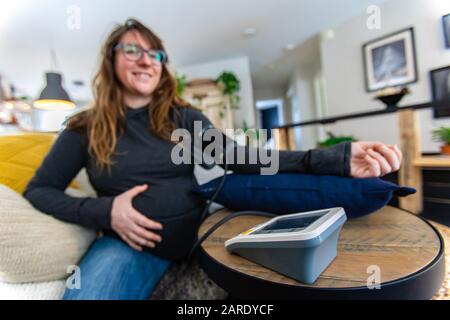 Ein selektiver Fokusschuss der Frau, die eine pränatale Gesundheitskontrolle hat, auf dem Sofa mit einem Sphygmomanometer, das den Blutdruck an der Arteria brachialis misst Stockfoto