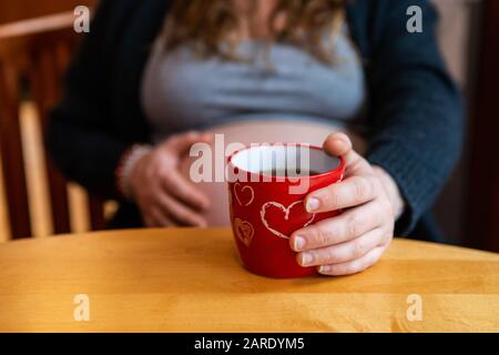 Eine Nahaufnahme einer schwangeren Frau, die am Valentinstag eine Tasse Tee genießt, serviert in roter Tasse mit Herzen, fühlt sich im Hintergrund unscharf bewegt Stockfoto