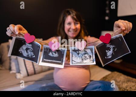 Eine selektive Fokusansicht einer lächelnden schwangeren Frau, die im Bett sitzt und Ultraschallaufnahmen zeigt, die einen gesunden Babyfötus in jeder Phase der Schwangerschaft zeigen Stockfoto