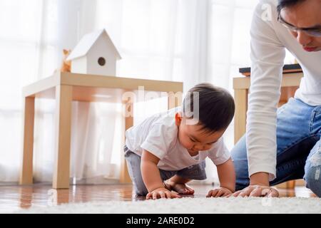 Ernster asiatischer kleiner Junge, der auf dem Boden krabbelt, während der junge Vater neben ihm im Wohnzimmer zu Hause sitzt Stockfoto