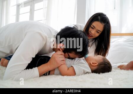 Die Familie des jungen asiatischen Vaters küsst seinen kleinen Jungen zärtlich mit seiner Frau auf den Bauch. Mann und Frau genießen ihre Zeit mit Kind Stockfoto