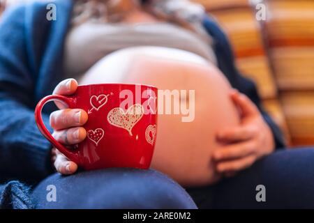 Ein kleiner selektiver Fokusschuss einer stark schwangeren Frau, die in der neuen roten valentinschale ein heißes Brühen genießt und die geschwollenen Bauch hält, die Babybewegung spüren Stockfoto