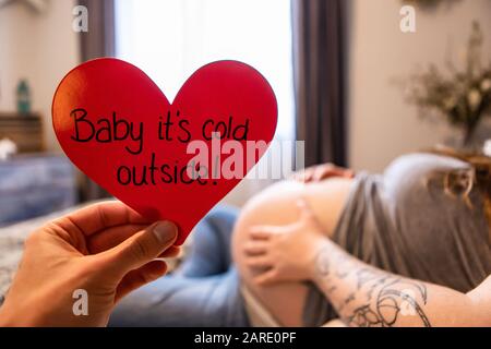 Eine selektive Fokusperspektive für die erste Person, in der eine Hand ein rotes Herz mit Botschaft Baby im Freien hält, während eine stark schwangere Frau im Bett ruht Stockfoto