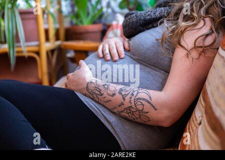 Ein Nahschuss einer stark schwangeren Frau im dritten Trimenon, der sich auf dem Sofa im Familienzimmer entspannt, Bewegungen spürt und mit den Händen in der Nähe der Arbeit tritt Stockfoto