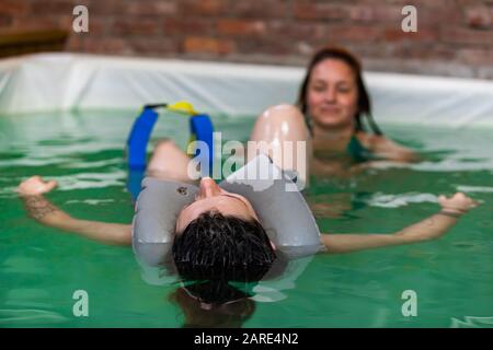 Junge Frau im Wasser auf dem Rücken mit Schwimmer um den Hals und Wassermassage vom Therapeuten im Schwimmbad während der Schwangerschaft Stockfoto