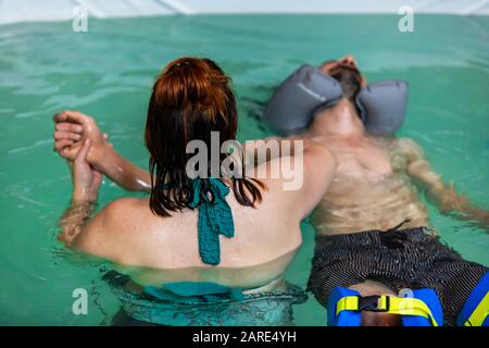 Rückansicht der Therapeutin mit Handwassermassage im Schwimmbad des jungen entspannten Mannes, mit Gummischwand um Hals und Bein schwebend Stockfoto