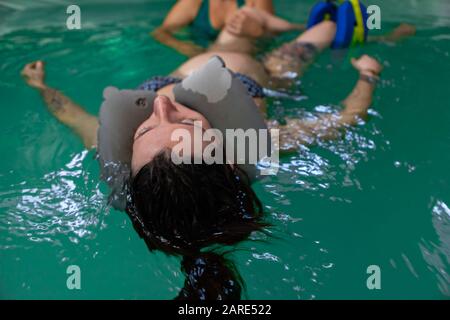 Nahaufnahme der schwangeren Frau mit Gummihalskissen, die auf dem Swimmingpool schwimmt, während sie sich entspannt und mit Hilfe von Trainer und Therapeut trainiert Stockfoto