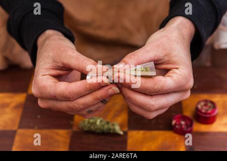 Nahaufnahme von Händen, die ein mit Cannabis gefülltes Rollpapier einklemmen, und beginnen, es in eine Verbindung zu rollen. Unkrautknospe und Mahlwerk auf der Tabelle unten. Stockfoto