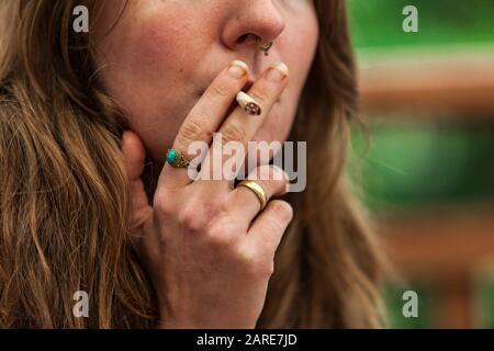 Closeup 3/4-Schuss des Gesichts der kaukasischen Frau mit handgerolltem Marihuana gefüllte Zigarette zwischen ihren Lippen. Cannabisgelenk wird im Freien geraucht. Stockfoto