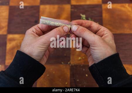 Oben Nahaufnahme von Fingern, die das Ende eines gemahlenen Cannabis gefüllten Rollpapiers quetschen, das es zu einem Gelenk über einem karierten Tisch im Hintergrund bildet. Stockfoto