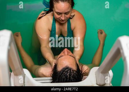 Hochwinkeliger Blick auf den jungen Mann, der sich an die Leiter des Schwimmbads lehnt und eine Kopfwassermassage im Schwimmbad von erfahrener Therapeutin erhält Stockfoto