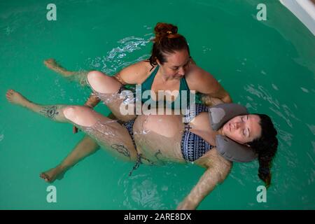 Hochwinkeliger Blick auf schwangere Frauen in Bikini und Tätowierung am Körper mit geschlossenen Augen, die Wassermassage erhalten. Von Therapeutin im Schwimmbad Stockfoto