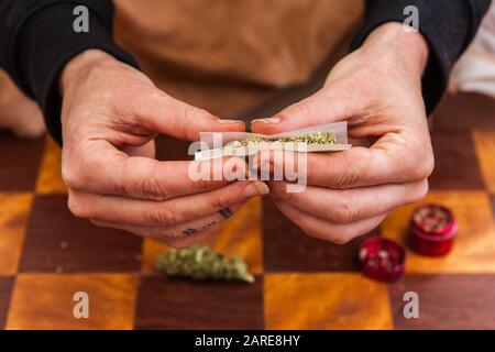 Nahaufnahme der Hände, die ein mit Cannabis gefülltes Rollpapier eingeklemmt haben, um es in ein Gelenk zu rollen. Unkrautknospe und Schleifer auf der Tabelle unten. Stockfoto