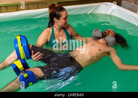 Junger Mann, der im Schwimmbad mit Nackenschwimmen und Beinauflage liegt, erhält Aqua-Behandlung für eine mit einer erfahrenen Trainerin Stockfoto