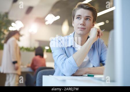 Portrait eines lächelnden jungen Mannes, der trostlos wegschaut, während er mit einem Laptop im Büro arbeitet, Platz für Kopien Stockfoto