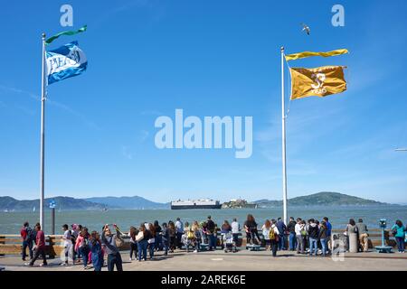 Am Ufer am Pier 39 versammelte sich eine Menge Besucher an einem sonnigen luftigen Tag mit Blick auf Alcatraz Island, als ein Frachtschiff vorbeisegelt. Stockfoto