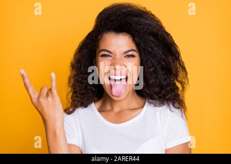 Foto des geschweiften, gewellten, aufgeregten, verrückten Mädchens, das mit den gehornenen Fingern die Zunge heraushält, ein in weißer Farbe isoliertes Rockschild mit einem T-Shirt Stockfoto