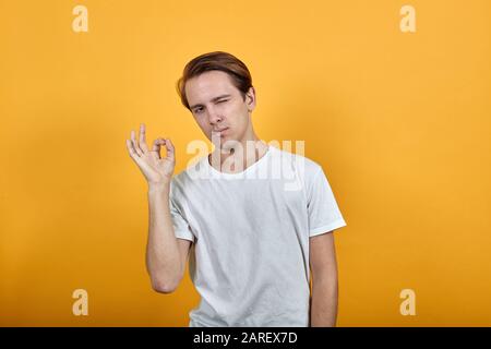 Ein junger Mann in weißem T-Shirt quetscht ein Auge und zeigt ein OK-Zeichen Stockfoto