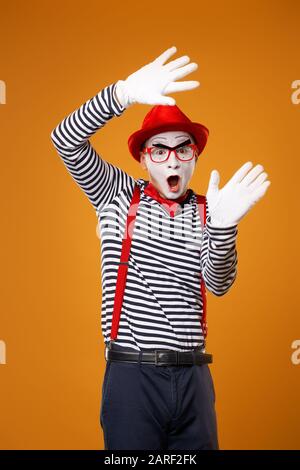 Überraschter MIME-Mann in Weste und roter Hut Isoliert auf leerem orangefarbenem Hintergrund Stockfoto