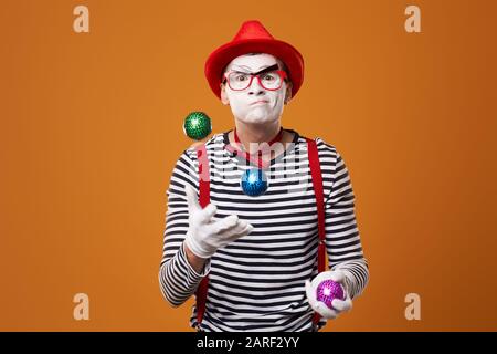 MIME man in Weste und Red hat jongliert mit bunten Bällen auf orange leerem Hintergrund Stockfoto