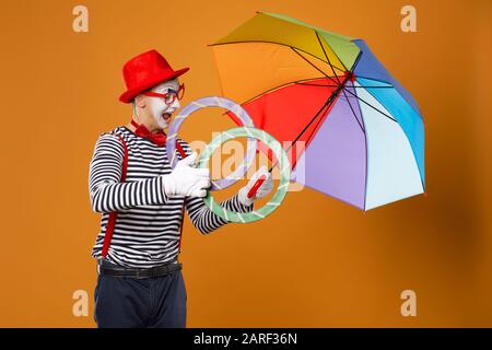 Fröhlicher MIME mit Ringen und mehrfarbigem Regenschirm auf leerem orangefarbenem Hintergrund im Studio Stockfoto