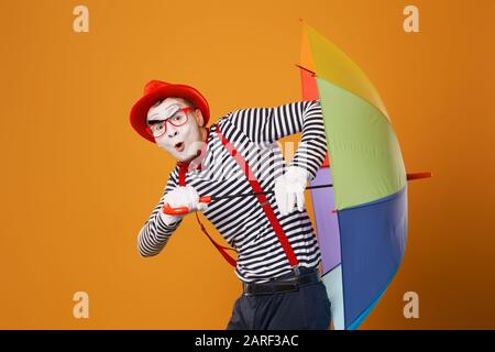 MIME man mit mehrfarbigem Regenschirm auf leerem orangefarbenem Hintergrund im Studio Stockfoto