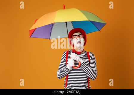 Überraschter MIME mit mehrfarbigem Regenschirm auf leerem orangefarbenem Hintergrund im Studio Stockfoto