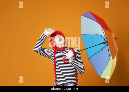 MIME man sucht mit mehrfarbigem Regenschirm, der auf leerem orangefarbenem Hintergrund im Studio isoliert ist Stockfoto