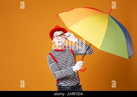 Trauriger MIME-Mann mit Blick auf die Kamera mit mehrfarbigem Regenschirm in der Hand isoliert auf leerem orangefarbenem Hintergrund im Studio Stockfoto