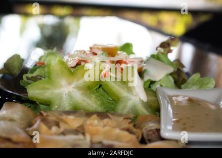 Dieses einzigartige Foto zeigt die Frühlingsrollen mit einem süß-sauren Dip im Vordergrund und einem Salat mit Sternfrüchten im Hintergrund. Stockfoto