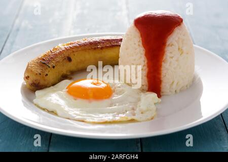 Arroz a la cubana Typisch kubanischer Reis mit gebratener Banane und gebratenem Ei auf blauem Holztisch. Stockfoto