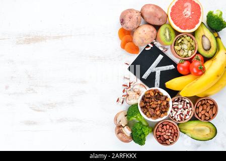 Lebensmittel, die natürliches Kalium enthalten. K: Kartoffeln, Pilze, Banane, Tomaten, Nüsse, Bohnen, Brokkoli, Avocados. Draufsicht. Auf weißem Holzhintergrund. Stockfoto