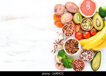 Lebensmittel, die natürliches Kalium enthalten. K: Kartoffeln, Pilze, Banane, Tomaten, Nüsse, Bohnen, Brokkoli, Avocados. Draufsicht. Auf weißem Holzhintergrund. Stockfoto