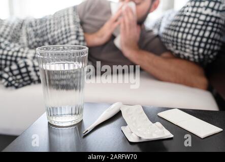 Medikamente, Thermometer und Glas Wasser auf Nachttisch gegen kranke männliche Person, die im Bett liegt und an Erkältung oder Grippeid leidet und seine Nase bläst Stockfoto