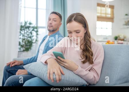 Tochter mit Smartphone, die vom Vater wegschaut, der auf dem Sofa sitzt. Stockfoto
