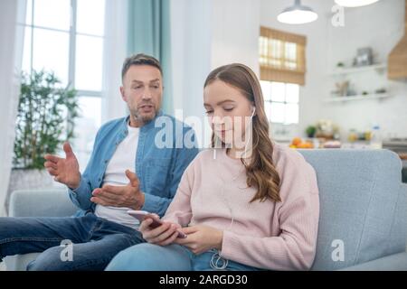 Tochter mit Smartphone, in Kopfhörern und Vater auf der Couch sitzen. Stockfoto