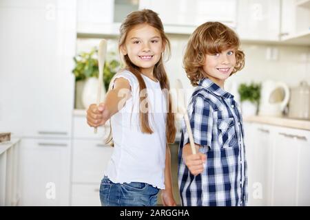 Zwei lächelnde Kinder halten Holzlöffel in der Küche in der Hand Stockfoto